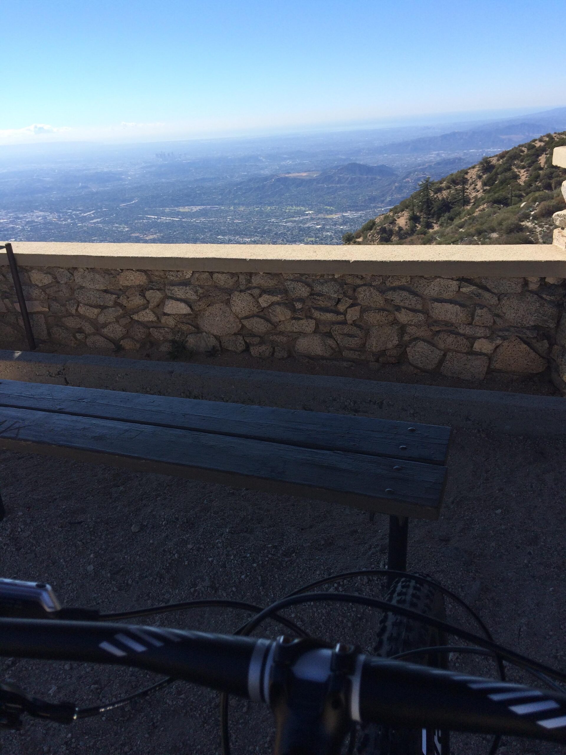 A panoramic view from a mountain overlook, showing a vast landscape with greenery and distant mountains under a clear blue sky. In the foreground, the handlebars of a mountain bike are visible, resting near a stone wall and a wooden bench. Idle Hour mountain bike trail.