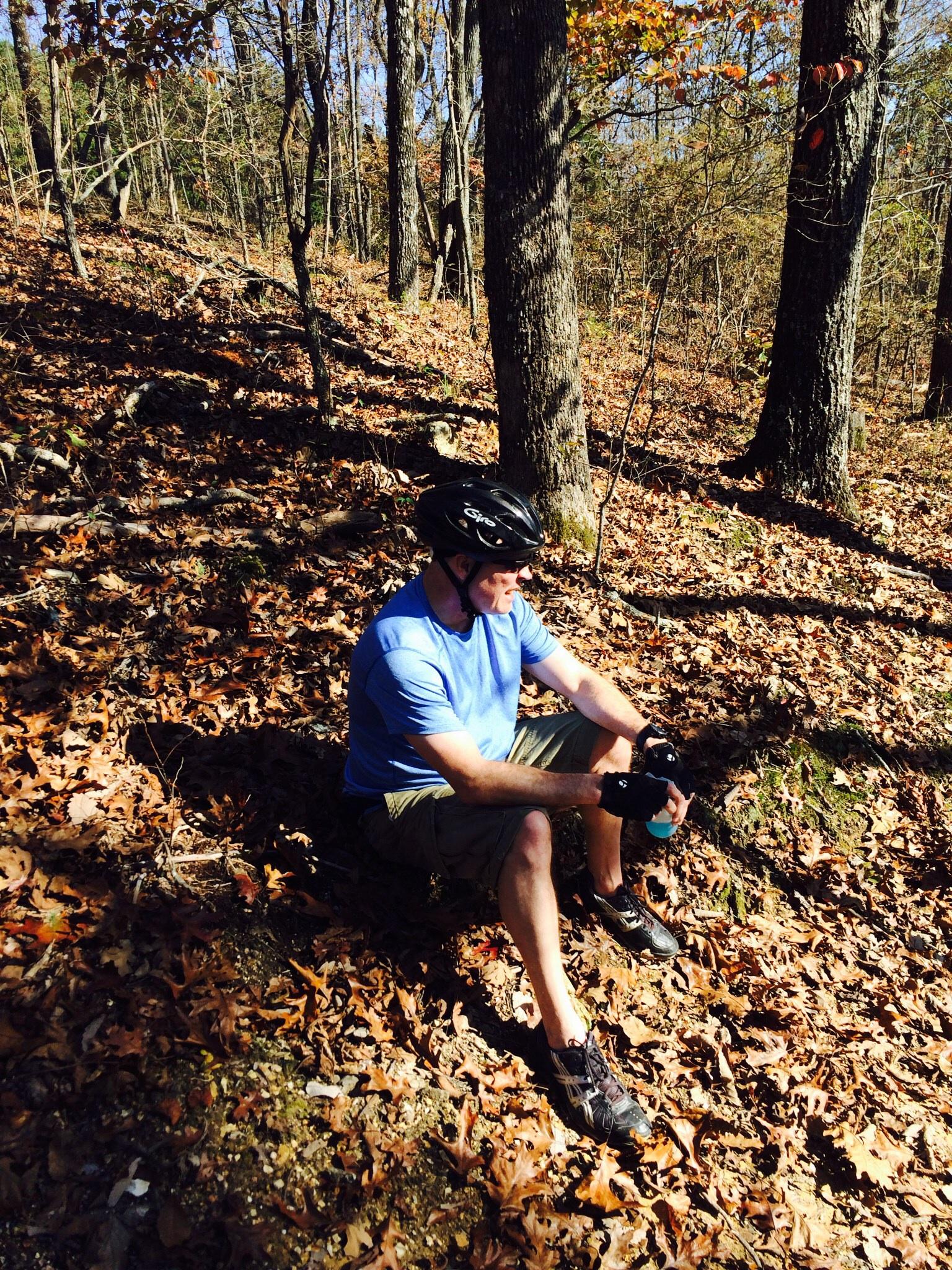 A person wearing a helmet and cycling gloves sits on the ground covered in autumn leaves, resting in a wooded area with trees in the background. They are holding a water bottle and wearing a blue shirt with shorts. The scene is illuminated by natural sunlight filtering through the trees. Enterprise South mountain bike trail.