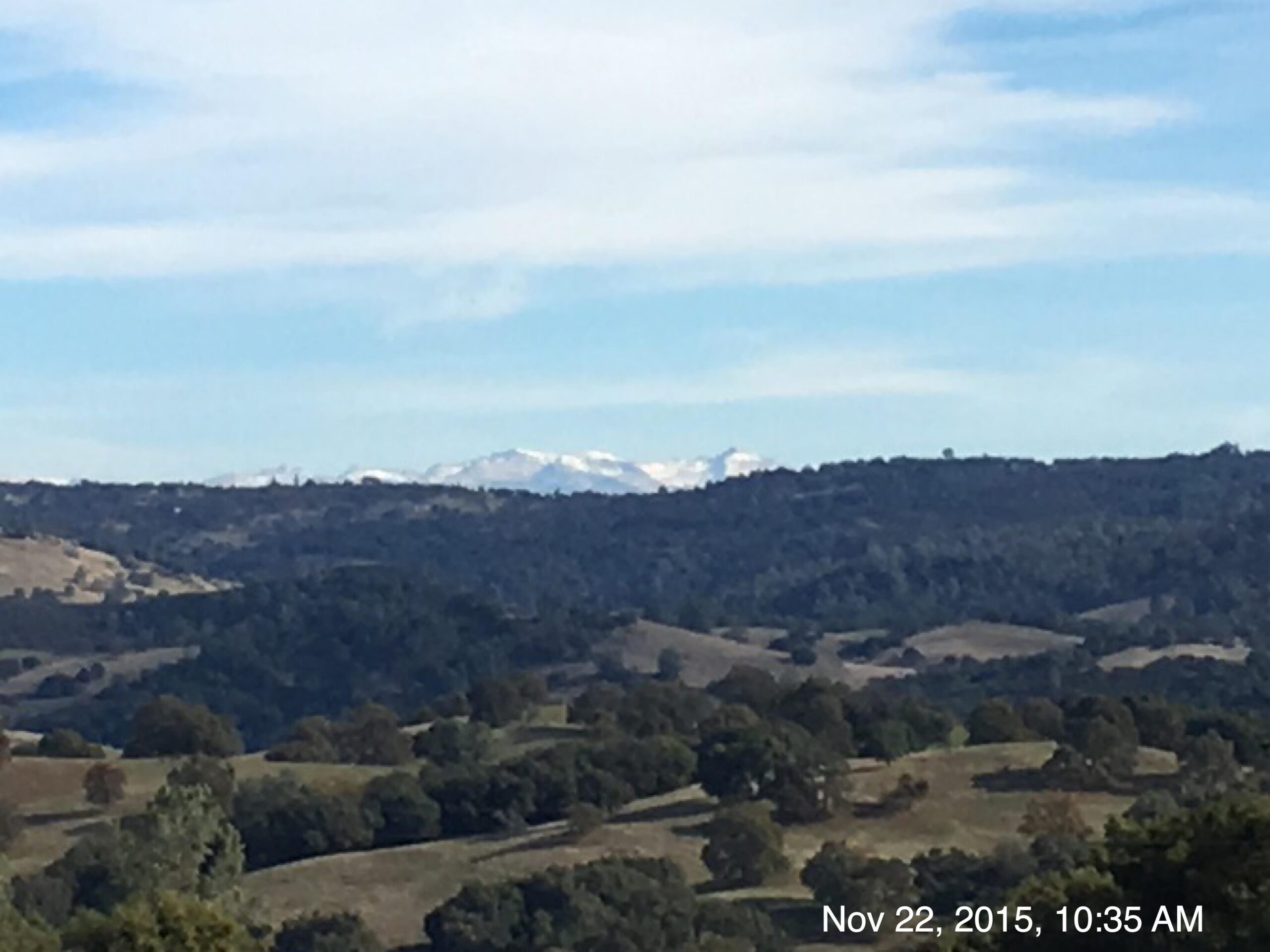 A panoramic view of rolling hills with sparse trees in the foreground, leading to a backdrop of distant snow-capped mountains under a clear blue sky. El Dorado Trail mountain bike trail.