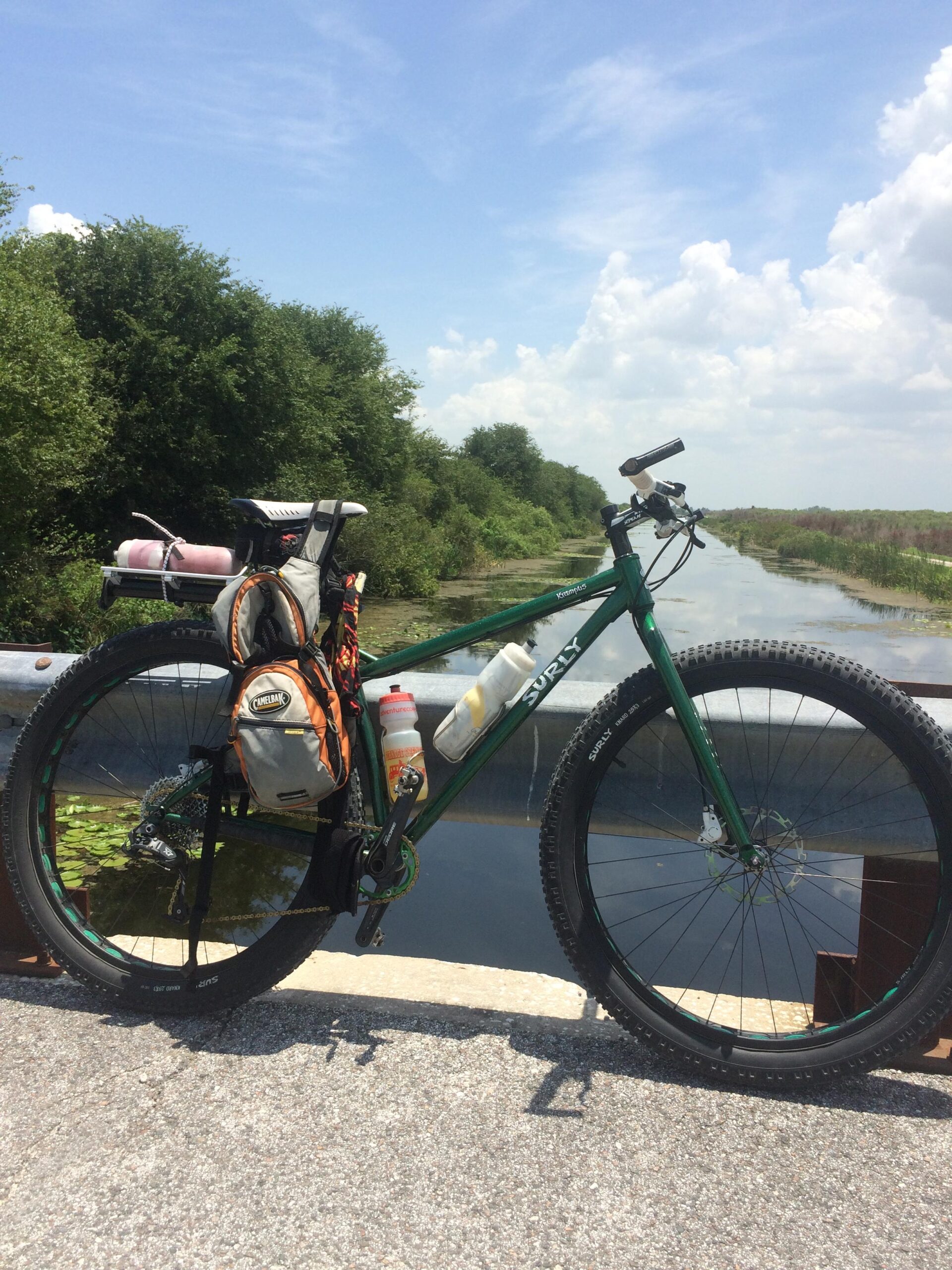 A green mountain bike is parked on a bridge beside a narrow, calm waterway, surrounded by lush greenery. The bike is equipped with a backpack and water bottles, indicating it is prepared for an outdoor adventure. The sky is bright with scattered clouds, and the scenery reflects a tranquil, sunny day. Lake Apopka Restoration Area mountain bike trail.