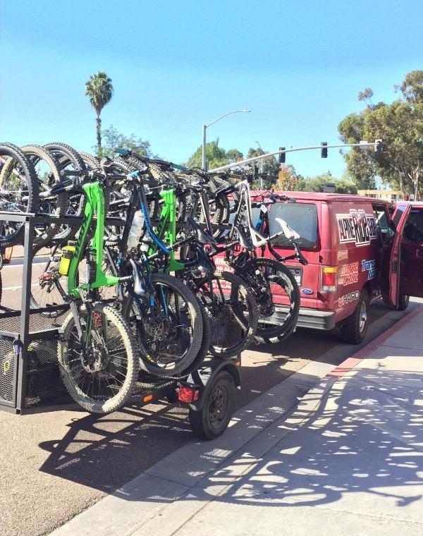 A trailer loaded with several mountain bikes is parked next to a red van with an open side door. The scene is bright and sunny, featuring palm trees in the background and a clear blue sky. The bikes include notable green and blue frames among a collection of black ones, all secured on the trailer.