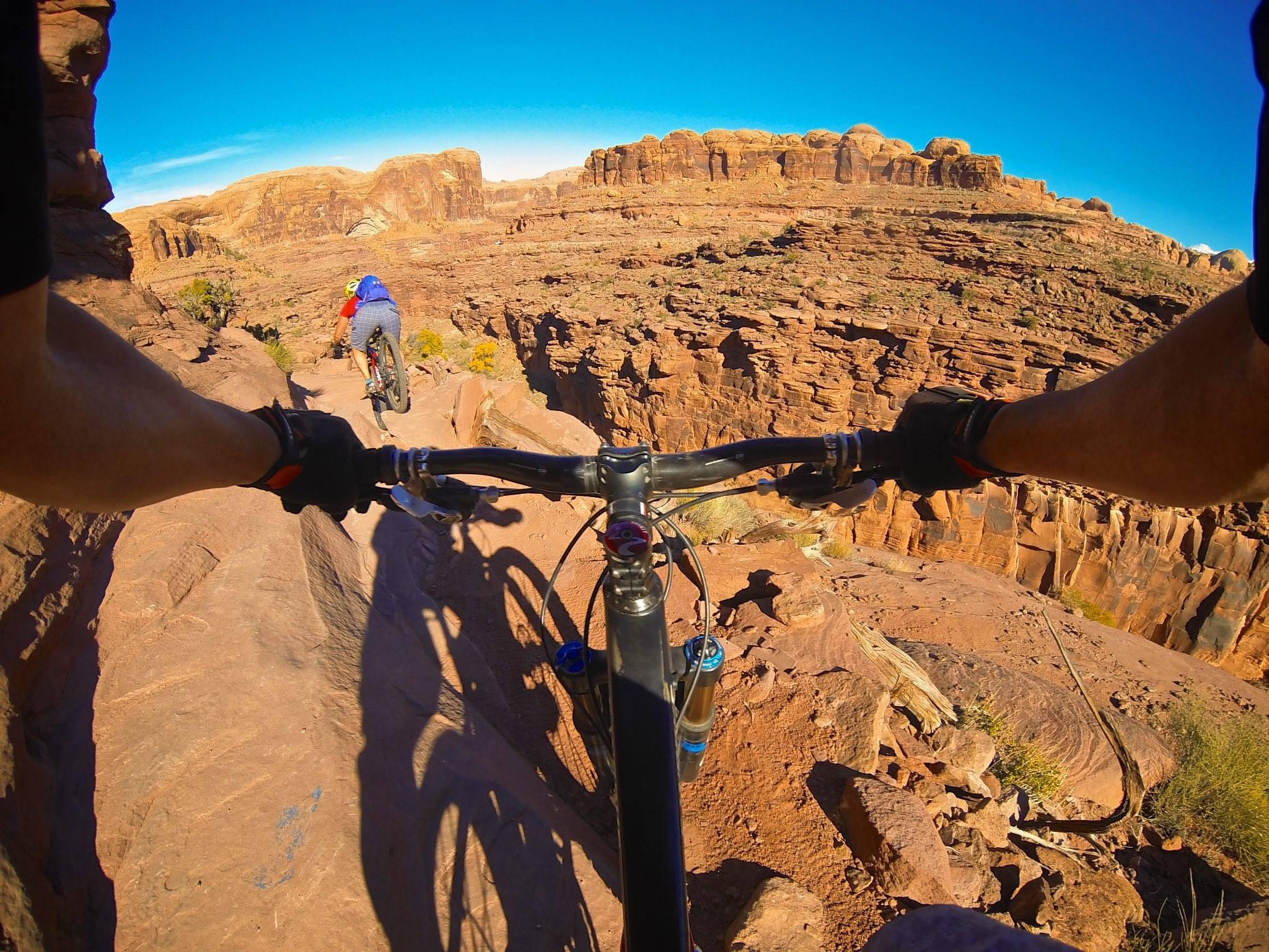 A view from a mountain bike rider's perspective, showcasing rocky terrain and a steep drop-off. Another cyclist in a blue shirt can be seen ahead, making their way along the narrow path against a backdrop of striking red rock formations and a clear blue sky. Captain Ahab mountain bike trail.