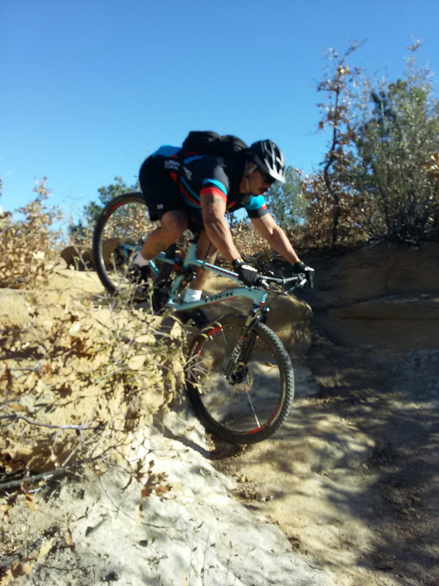 A person wearing a helmet and cycling gear is navigating a rocky terrain on a mountain bike. The cyclist leans forward as they ride over a rugged surface surrounded by sparse vegetation and blue sky. Palmer Park mountain bike trail.