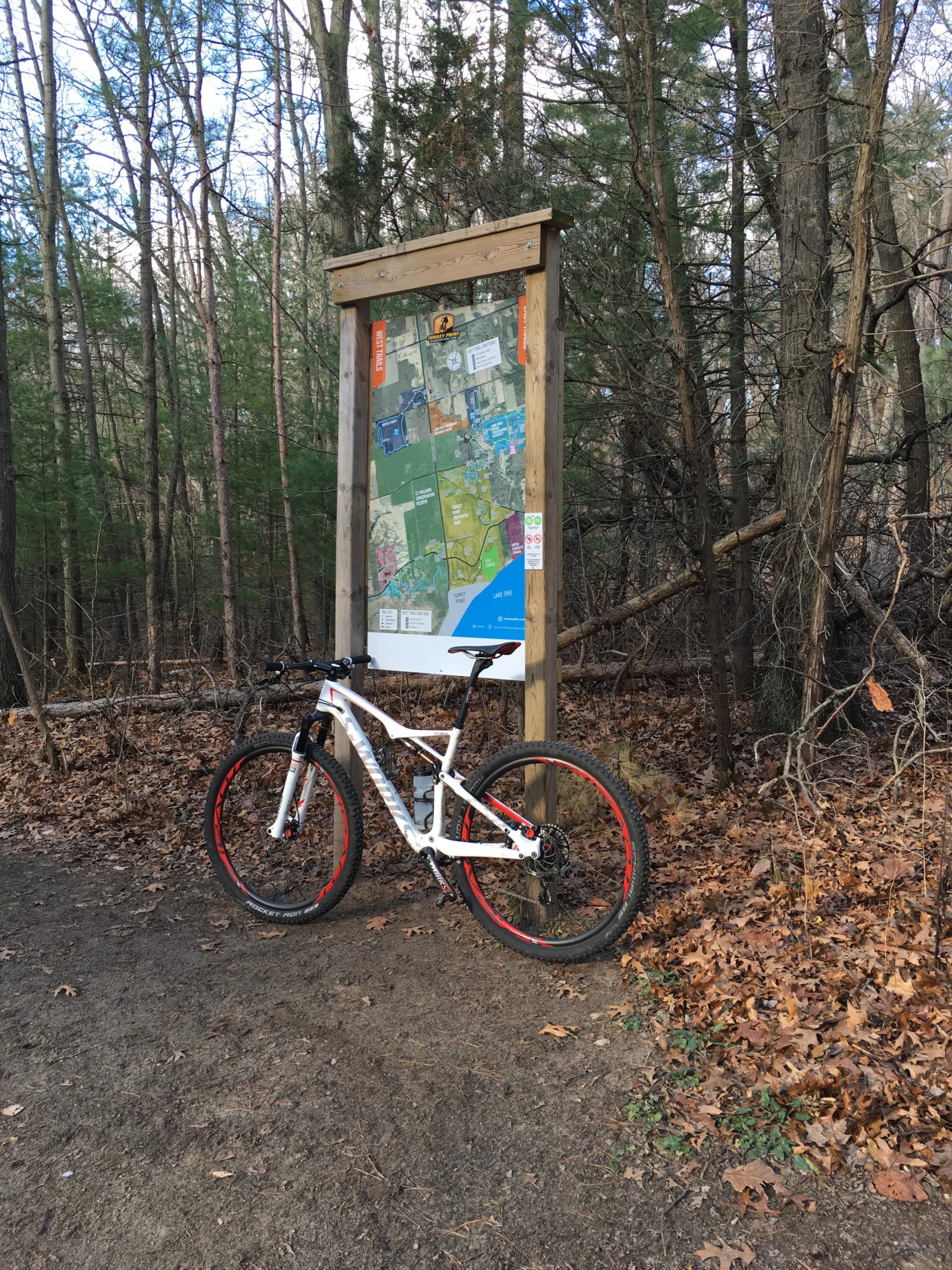 A mountain bike stands beside a wooden trail map sign in a forested area. The ground is covered with leaves, and trees surround the scene, indicating a natural outdoor environment. Turkey Point Provincial Park mountain bike trail.