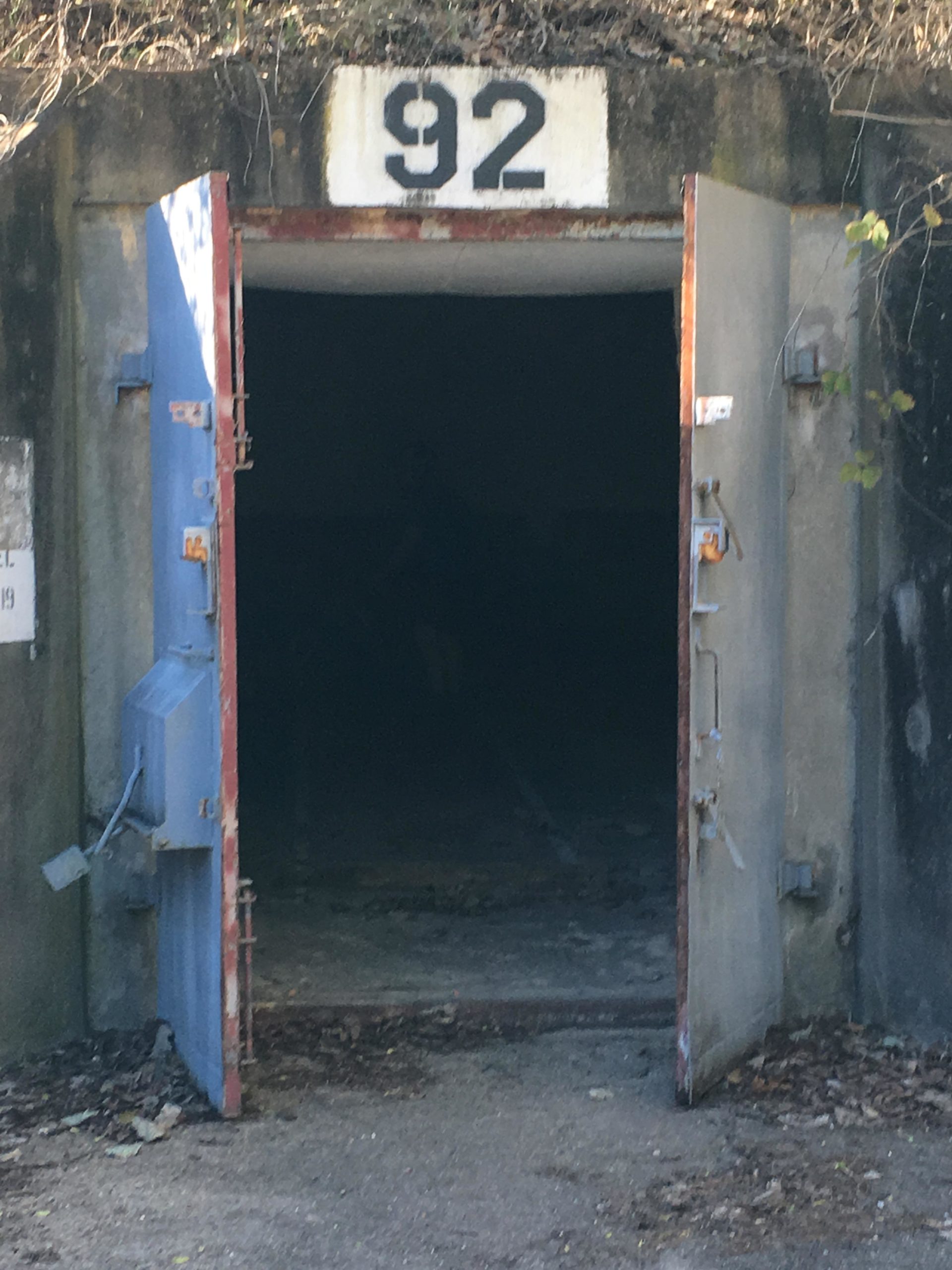 An entrance to a dark underground bunker, labeled "92" at the top, with two doors wide open and surrounded by overgrown vegetation and leaves on the ground. Enterprise South mountain bike trail.