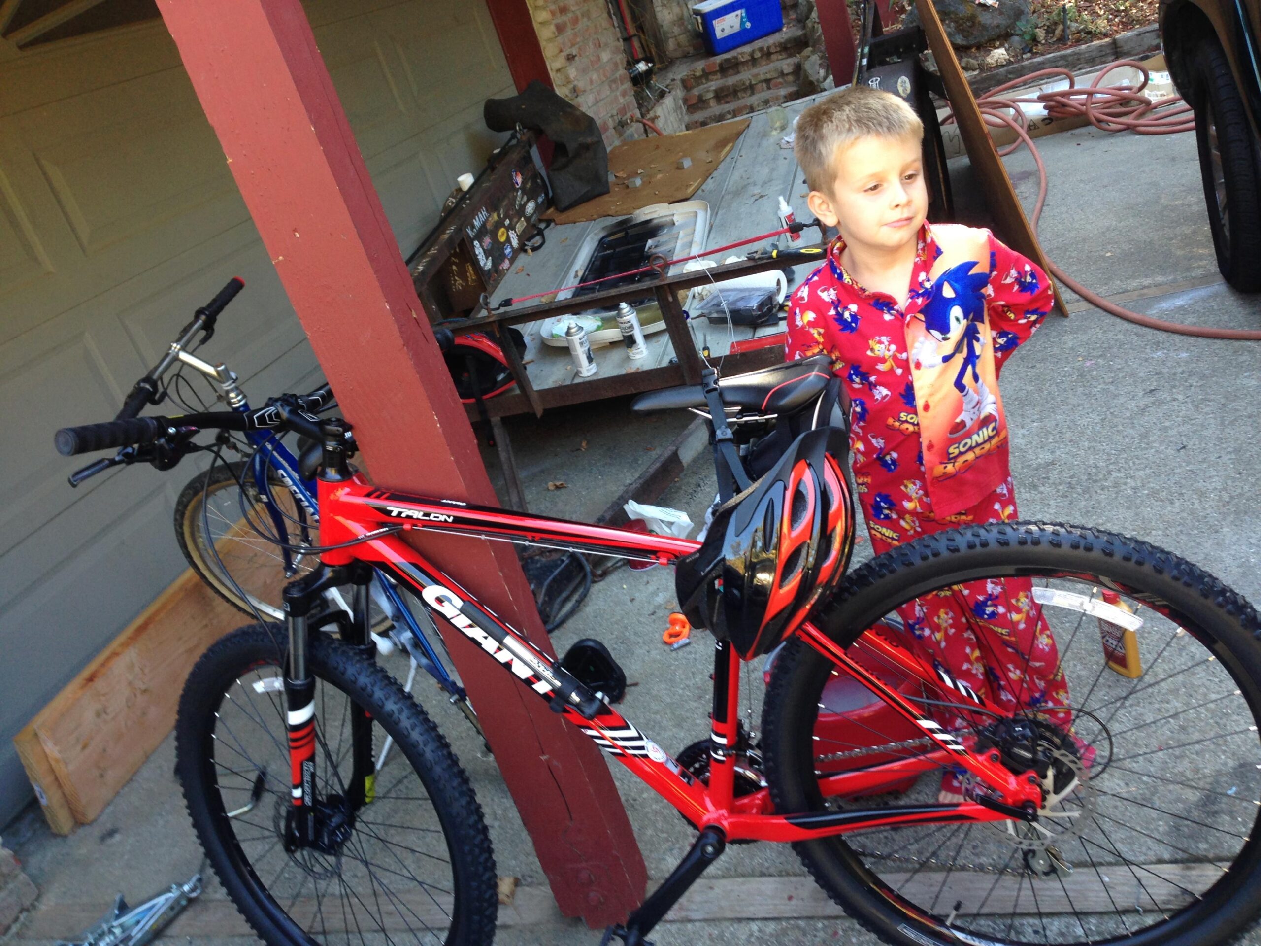 Giant Talon 27.5: A young boy in colorful pajamas stands next to a red mountain bike. He is holding a drink and looking thoughtfully off to the side. The background features a garage setting with tools and other bikes visible.