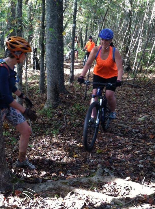 Specialized Rumor: Two women mountain biking on a wooded trail. One woman is riding a bike along the path, wearing an orange tank top and a helmet, while the other woman stands nearby, wearing a blue shirt and looking towards the cyclist. In the background, more cyclists can be seen riding along the trail surrounded by trees and fallen leaves.