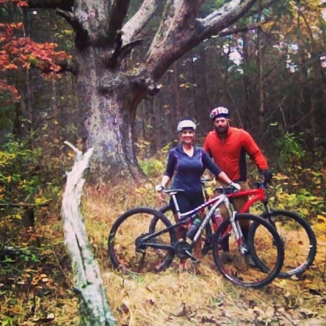 Two mountain bikers pose together with their bikes in a wooded area. The backdrop features a large, gnarled tree, while the ground is covered with autumn leaves. The riders are wearing helmets and casual biking gear, and they appear to be enjoying the outdoor setting. Binder Lake mountain bike trail.