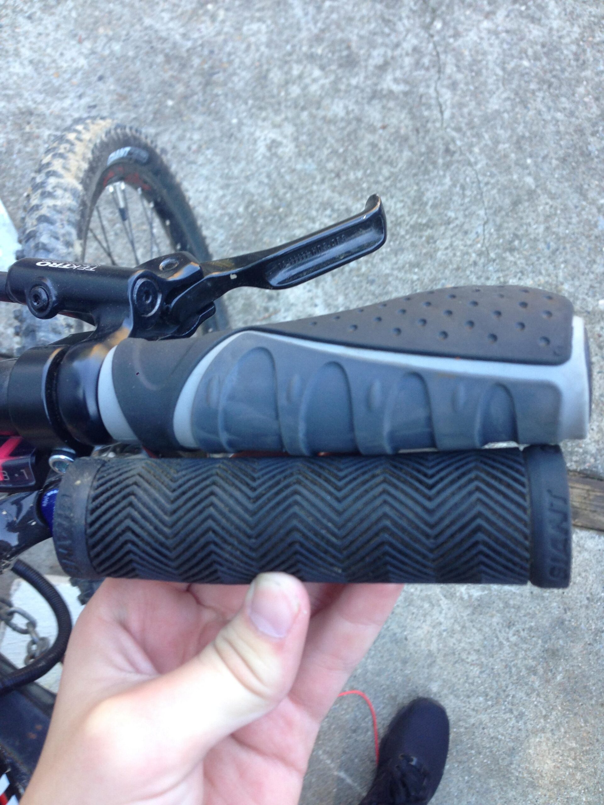 Giant Talon 27.5: A close-up view of two bicycle handlebar grips held in a person's hand. The top grip features a textured surface with a gray and black color scheme, while the bottom grip is black with a herringbone pattern. In the background, part of a mountain bike can be seen, along with a gravel surface.