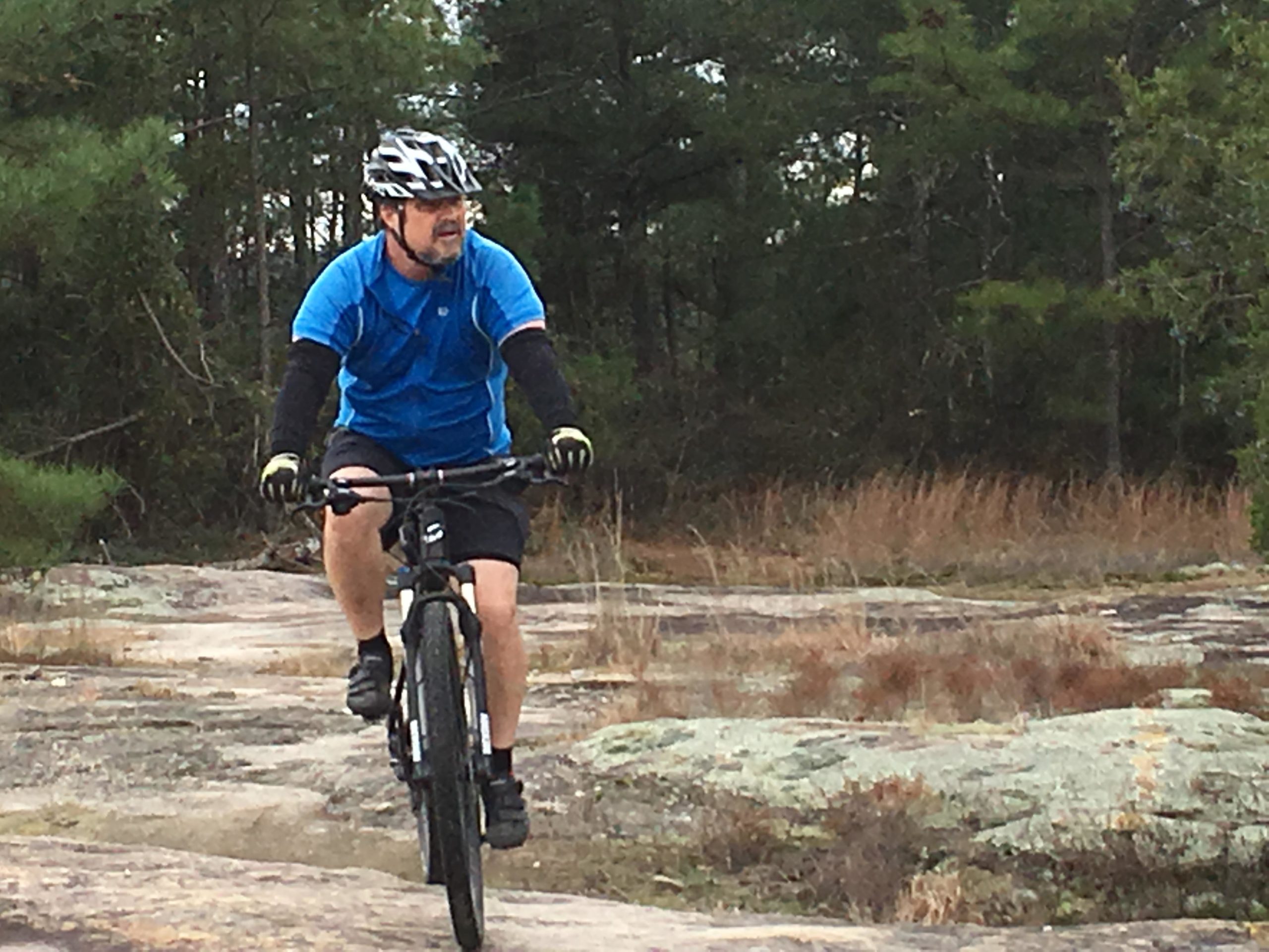A person wearing a blue shirt, black shorts, and a helmet rides a mountain bike on rocky terrain surrounded by trees and tall grass. Georgia International Horse Park mountain bike trail.