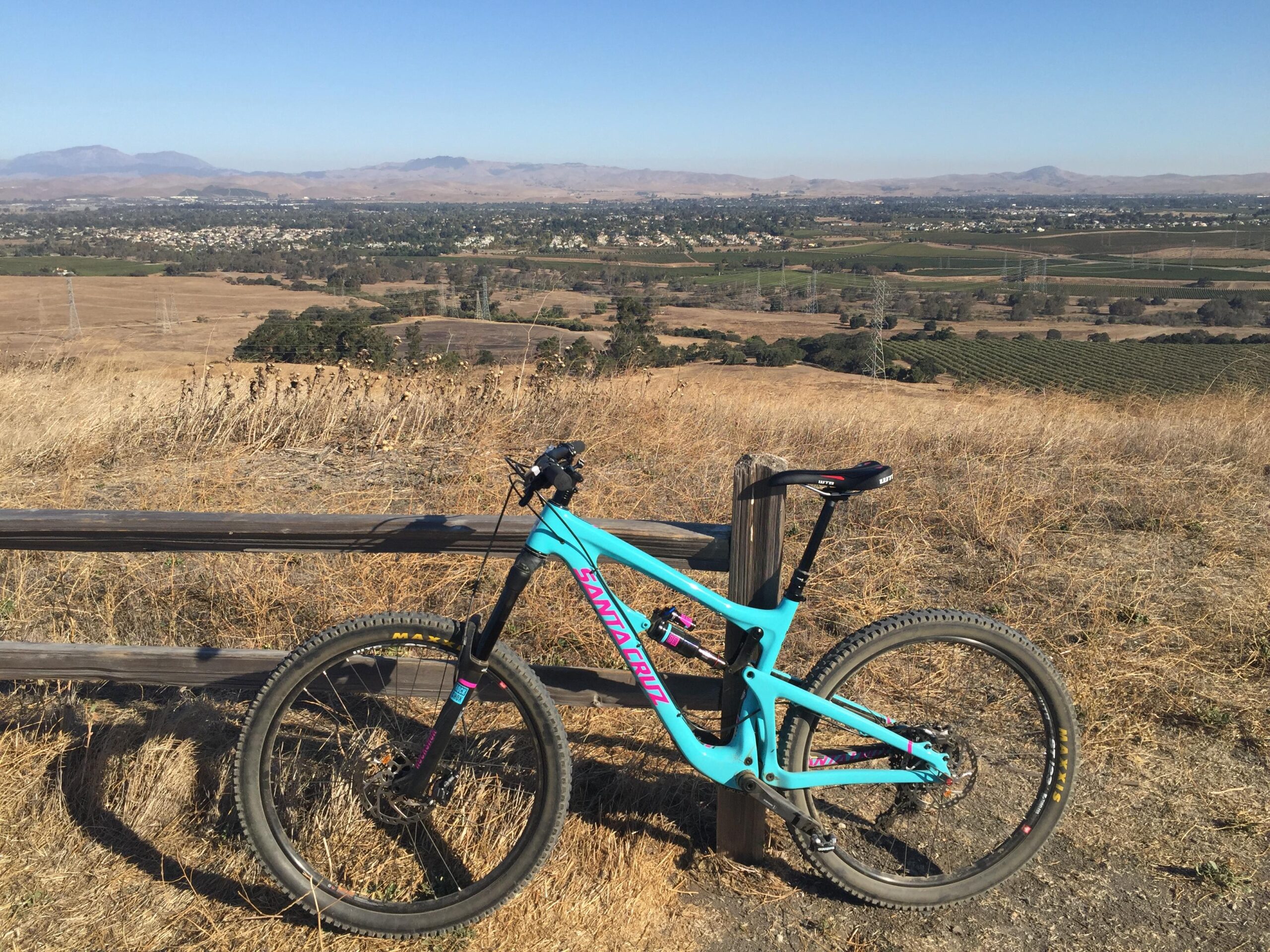 Santa Cruz Nomad: A bright blue mountain bike is leaning against a wooden fence, overlooking a wide valley filled with dry grass and patches of green fields. In the background, hills and mountains stretch into the distance under a clear blue sky. The scene captures a peaceful outdoor landscape, ideal for cycling and enjoying nature.