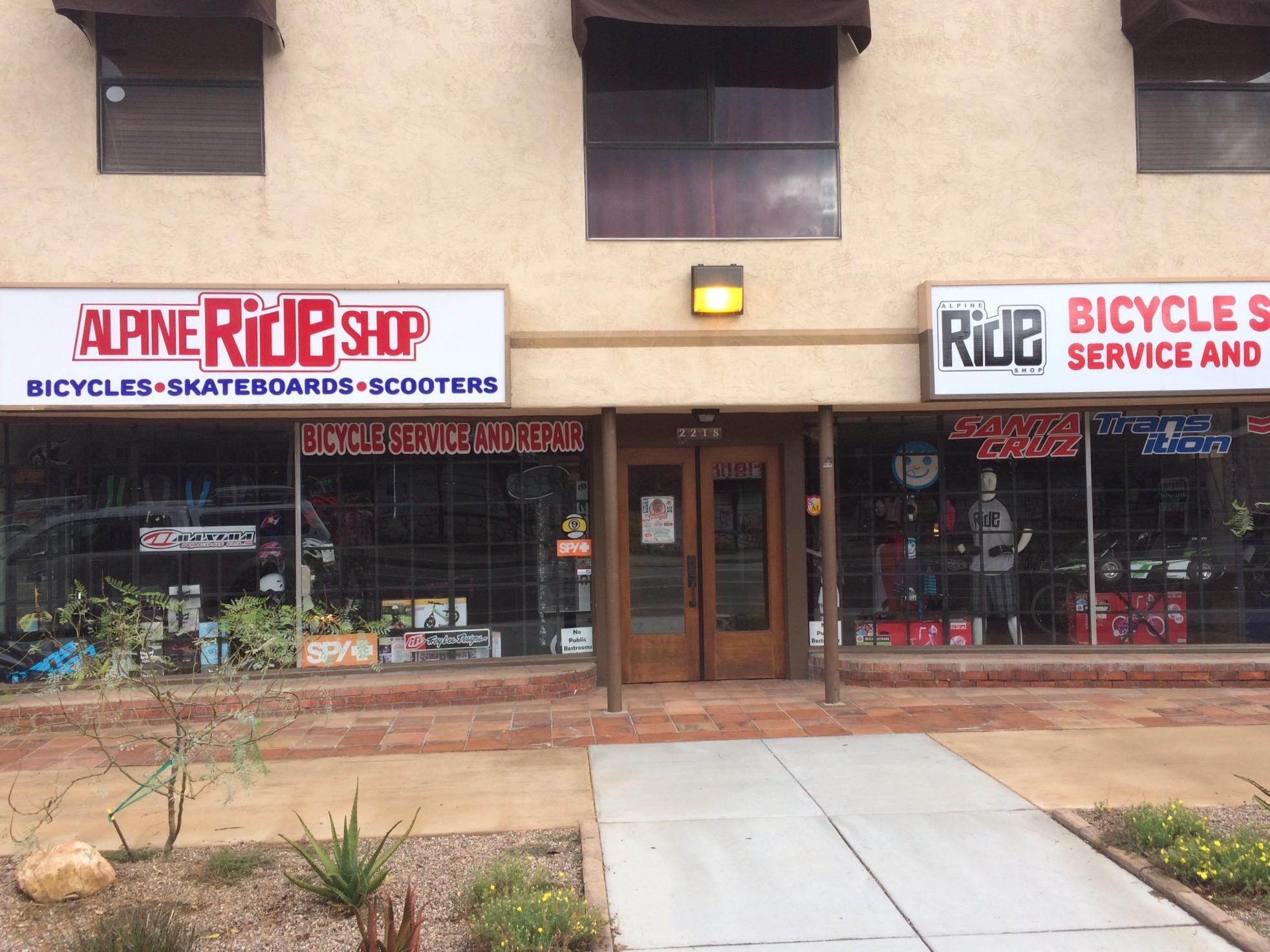 Image of a storefront featuring two signs for "Alpine Ride Shop," highlighting bicycles, skateboards, and scooters. The window displays showcase various bicycle parts and gear, with a focus on bicycle service and repair. The exterior has a desert landscape with some low plants and a paved sidewalk.