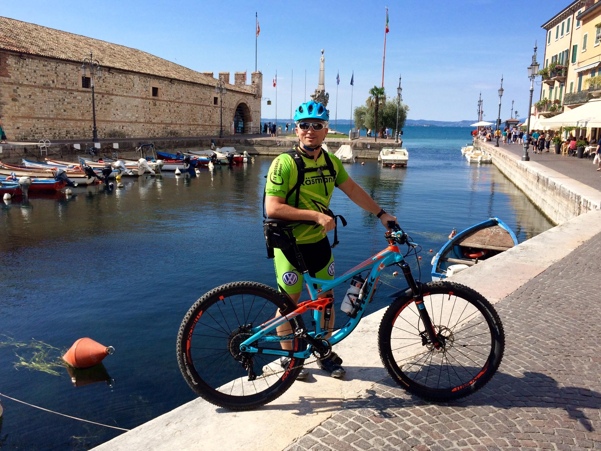 Trek Remedy 9: A cyclist in a green and black athletic outfit, wearing a blue helmet, stands next to a mountain bike on a cobblestone path beside a serene harbor. In the background, small boats are moored at the water's edge, with a historic stone building and colorful shops lining the waterfront. Flags flutter in the gentle breeze, and the scene is bathed in bright sunlight, evoking a lively summer ambiance.