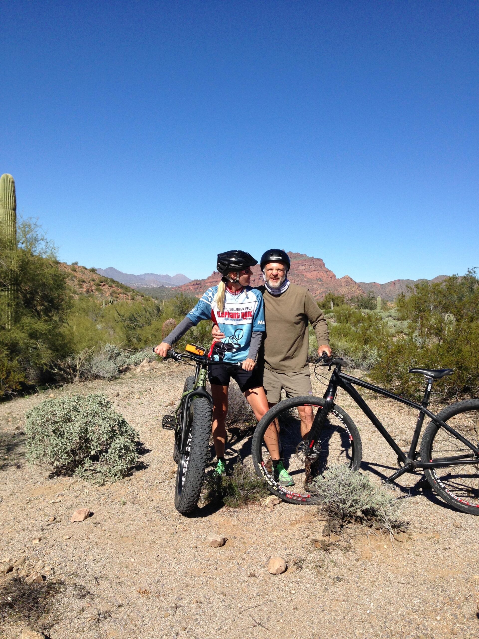 A young person and an older man are posing for a photo while standing beside their mountain bikes in a desert landscape. They are both wearing helmets and casual cycling attire, with a backdrop of rocky mountains and a clear blue sky. Cacti and desert vegetation are visible around them, indicating an outdoor biking adventure. Hawes Loop mountain bike trail.