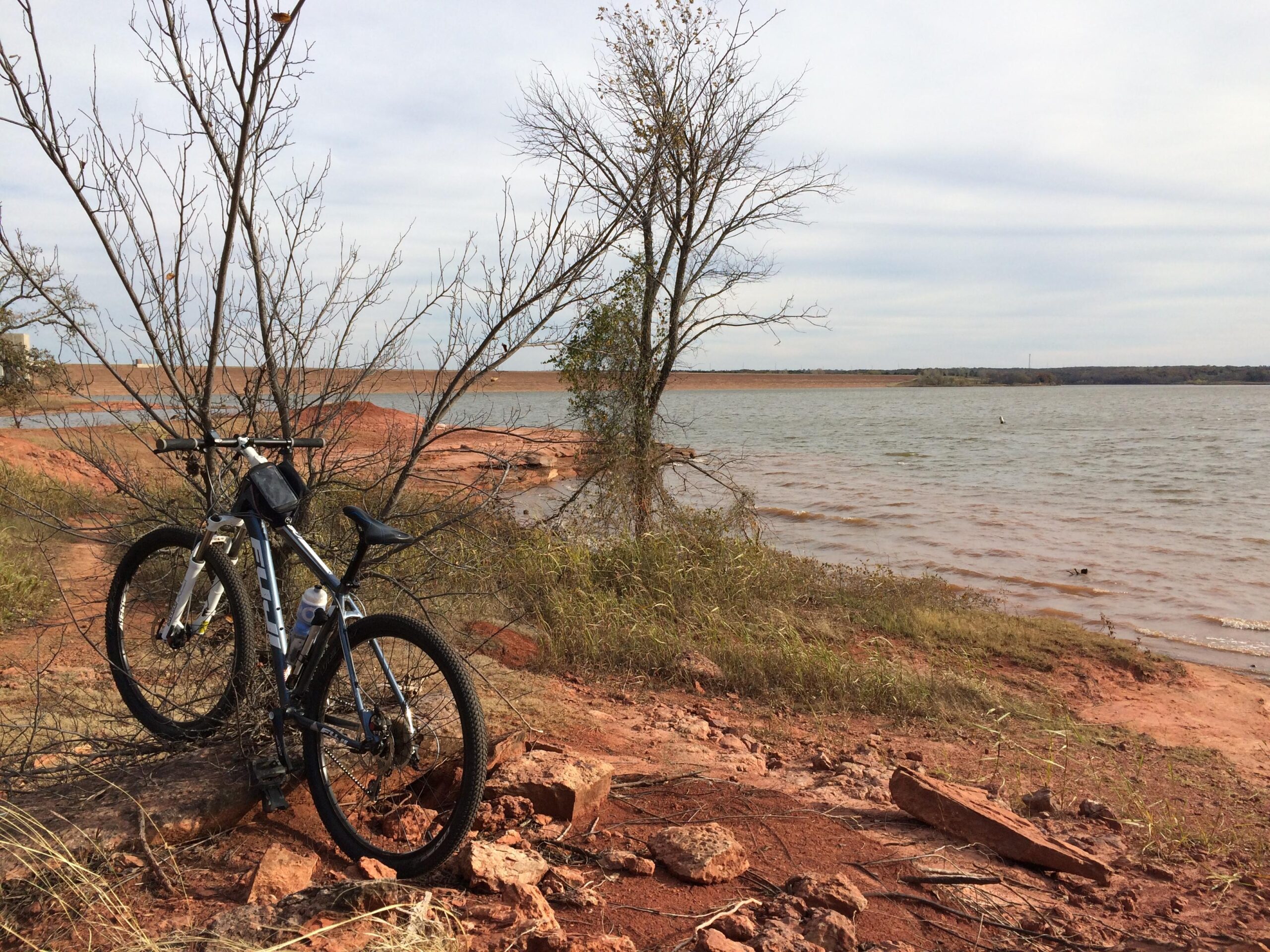 Fuji Nevada 29 1.7: A mountain bike rests near a lakeshore, surrounded by sparse trees and rocky terrain. The water's edge reflects a cloudy sky, with distant hills visible across the lake. The landscape features dry grass and red soil, creating a serene outdoor scene.