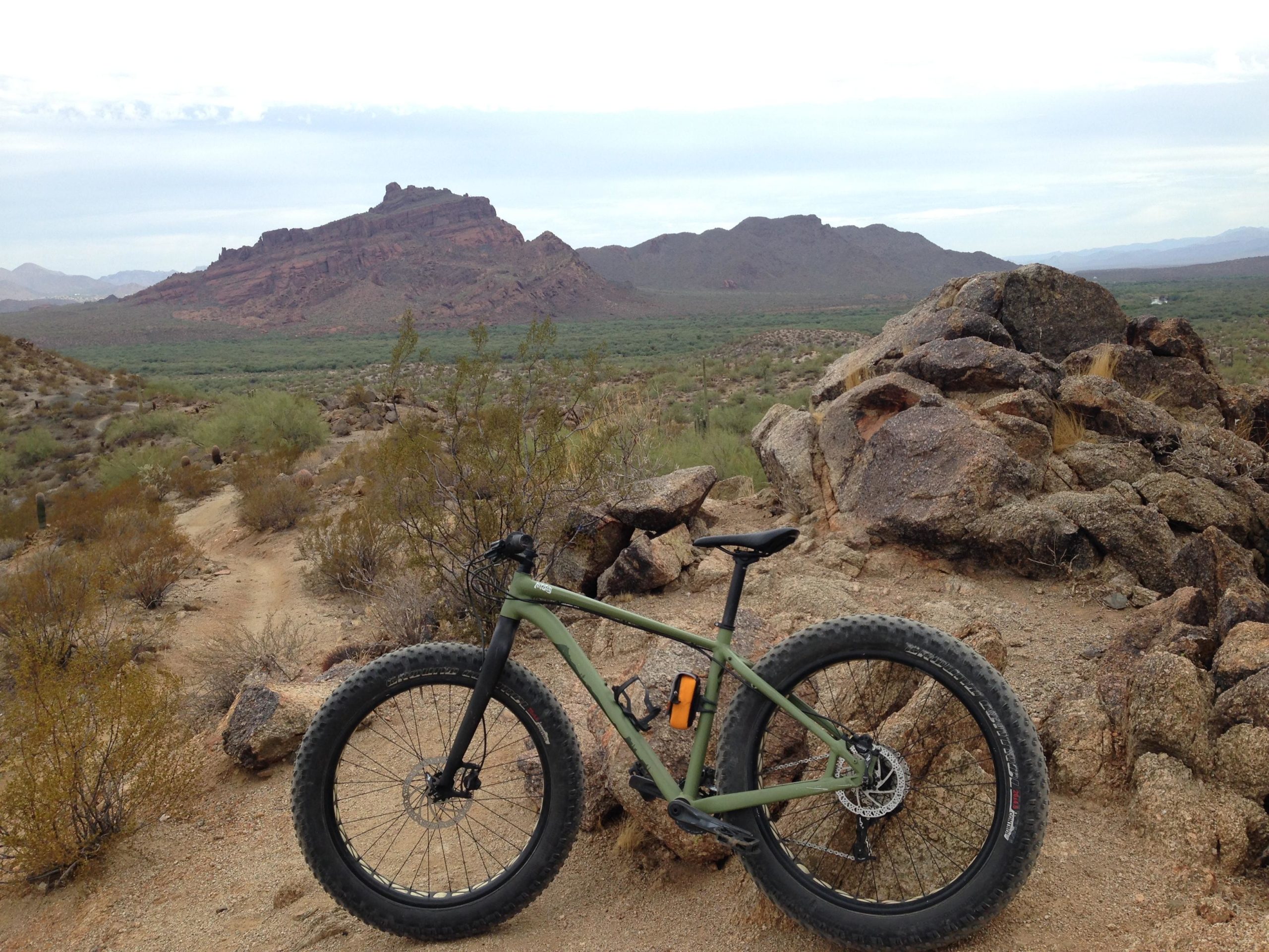 A rugged mountain bike rests on rocky terrain, with a scenic view of a mountainous landscape in the background. The area is characterized by desert vegetation and a winding dirt path that leads through the arid environment under a cloudy sky. Hawes Loop mountain bike trail.