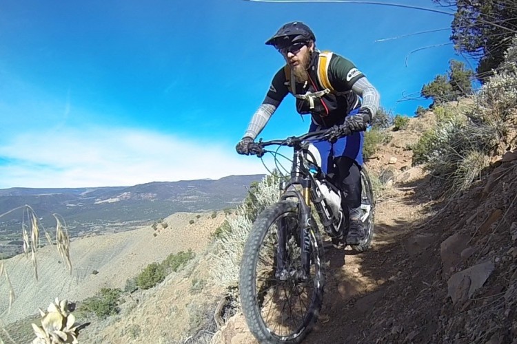 A mountain biker riding on a rocky, elevated trail surrounded by shrubs and trees, with a clear blue sky in the background. The cyclist is wearing a helmet and protective gear, showcasing a focused expression as they navigate the terrain.