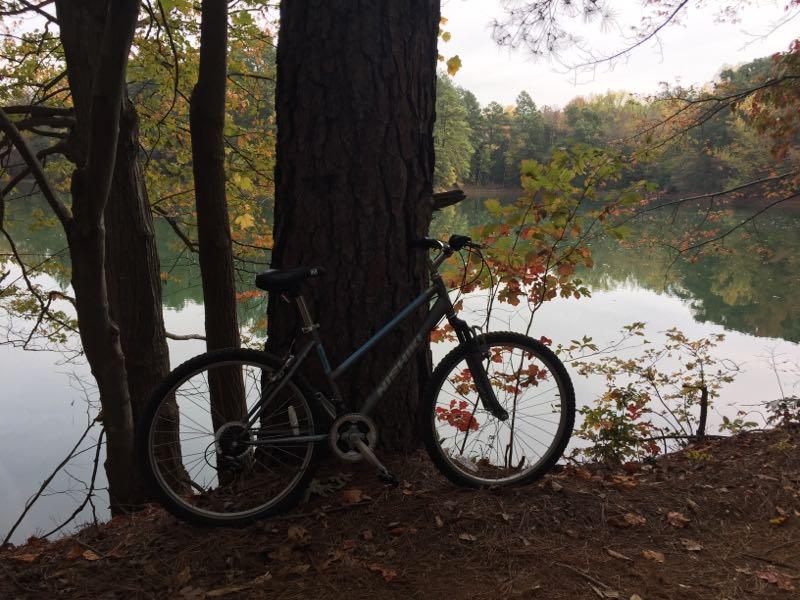 A mountain bike resting against a tree near a calm lake surrounded by trees displaying fall foliage. The scene captures a peaceful outdoor setting with reflections in the water. USNWC mountain bike trail.