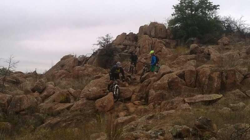 Four mountain bikers navigate a rocky terrain with scattered boulders and sparse vegetation under a cloudy sky. Lake Lawtonka Trails mountain bike trail.