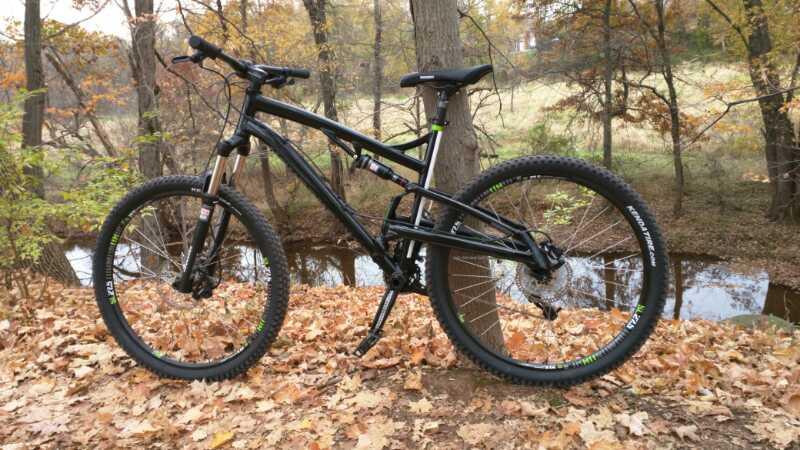 A black mountain bike leaning against a tree, surrounded by autumn leaves, with a small creek visible in the background and colorful foliage in a serene outdoor setting. Six Mile Run mountain bike trail.