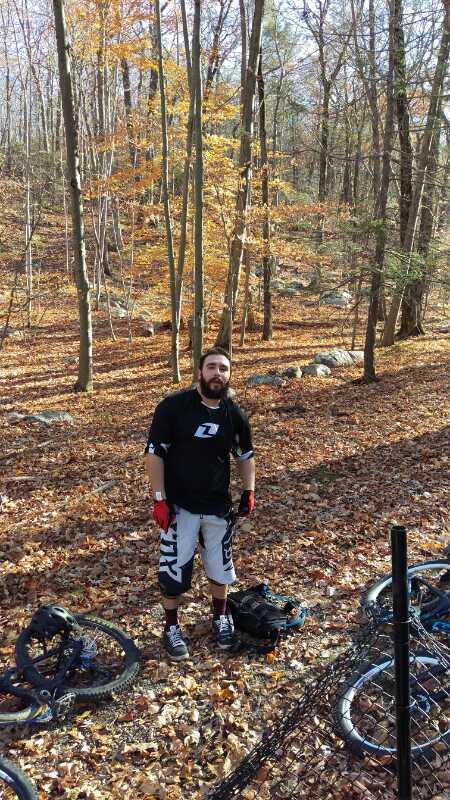 A man in biking gear stands in a forested area covered with autumn leaves, surrounded by trees with golden foliage. Several bicycles are parked nearby, and the man appears to be enjoying a break from mountain biking. Long Pond Ironworks State Park mountain bike trail.