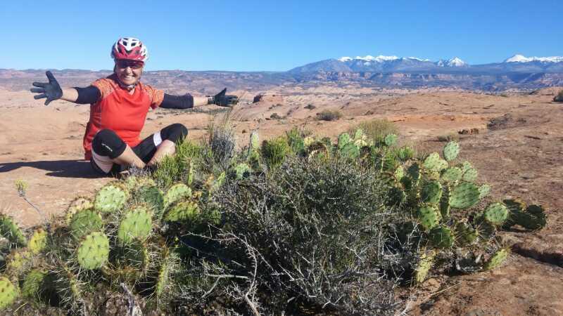 A person in a red cycling shirt and helmet is smiling and sitting among desert vegetation, including a cactus. They are positioned on a rocky landscape with mountains in the background under a clear blue sky. The scene conveys a sense of adventure and exploration in an arid environment. Slickrock mountain bike trail.