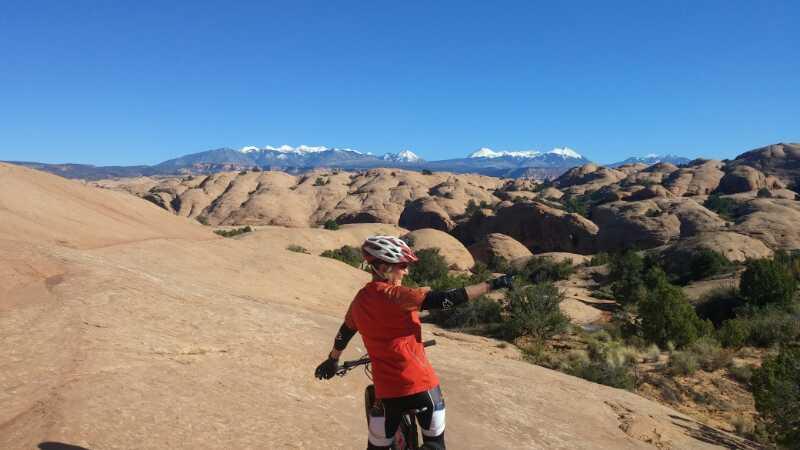 A person in a red shirt and cycling gear stands on a rocky landscape, pointing toward a distant mountain range with snow-capped peaks under a clear blue sky. The terrain consists of large, smooth rocks and scattered vegetation. The scene conveys a sense of adventure and exploration in an outdoor setting. Slickrock mountain bike trail.