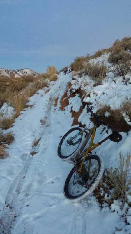 A mountain bike leaning against a snow-covered dirt trail surrounded by sparse vegetation and rolling hills under a clear sky. Bad Idea! mountain bike trail.