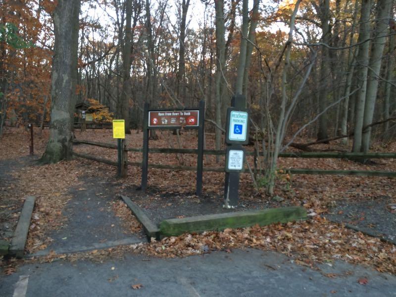 A wooded entrance to a trail, featuring a sign with trail rules and information. The ground is covered with fallen leaves, and there are benches and a wooden fence along the path. A parking area with signs indicating accessible parking is also visible. The scene is set in autumn with trees showing various shades of orange and brown. Hartshorne Woods Park mountain bike trail.