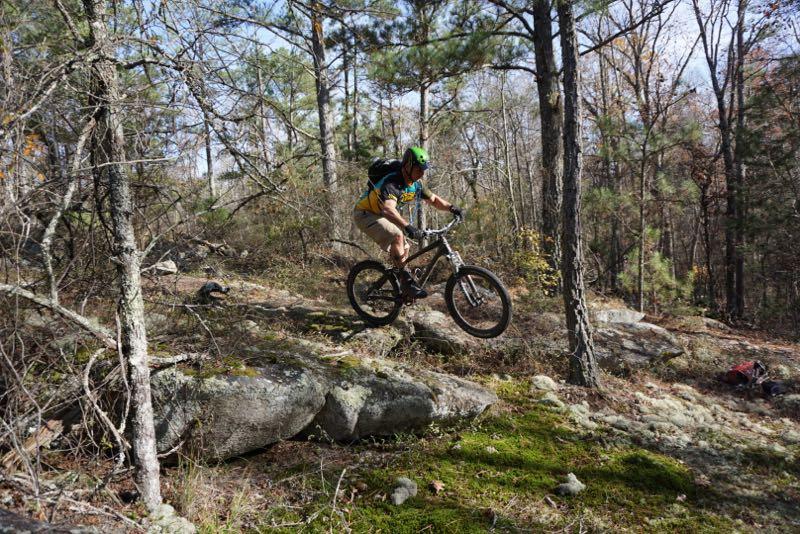 A mountain biker in a green helmet and colorful shirt jumps off a rocky path in a forested area. Surrounding trees and undergrowth create a natural backdrop, showcasing the rider's dynamic movement on the bike. Clinton Nature Preserve mountain bike trail.