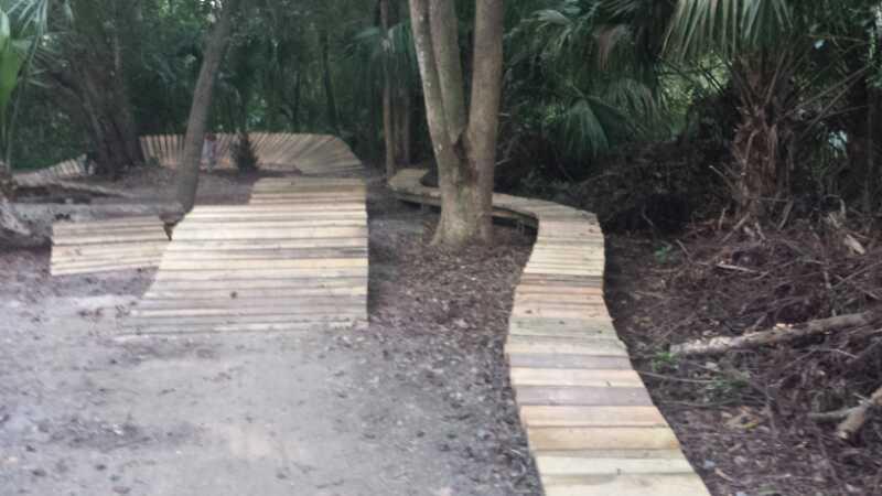A winding wooden pathway meanders through a forested area, surrounded by lush greenery and trees, with patches of earth and foliage visible along the sides. Mount Dora Trail mountain bike trail.