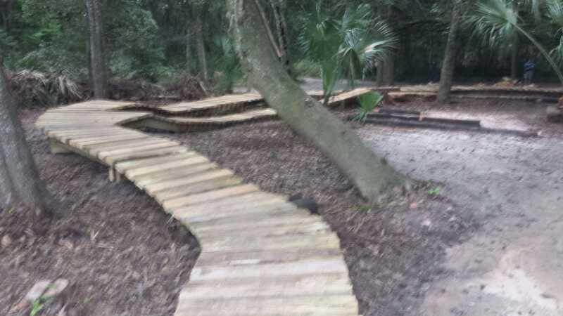 Curved wooden walkway winding through a wooded area, surrounded by trees and underbrush. Mount Dora Trail mountain bike trail.