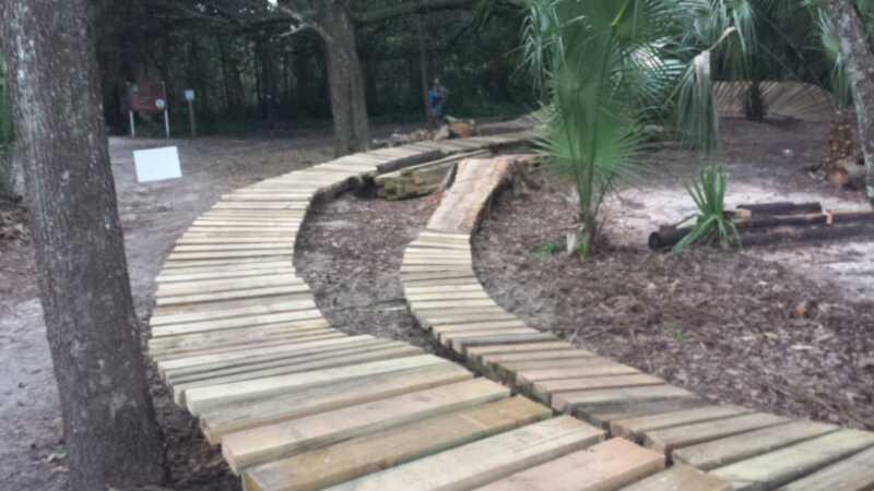A winding wooden pathway through a natural setting, surrounded by trees and vegetation. The path is made of planks and curves gently, leading into a forested area. There is a signpost in the background and a glimpse of a person at a distance. Mount Dora Trail mountain bike trail.