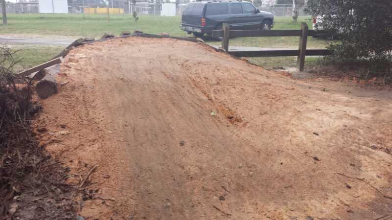 A sandy, unpaved path curves gently, with a dirt slope on one side and a wooden fence in the background. A parked black vehicle is visible in the distance, surrounded by greenery. The area appears to be outdoors, possibly in a park or recreational space. Mount Dora Trail mountain bike trail.