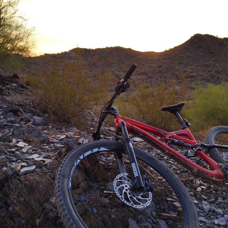 A red mountain bike resting on rocky terrain with a backdrop of hills and a sunset sky. The scene captures the natural beauty of an outdoor biking trail, showcasing the rugged landscape and vegetation. Trail #100 mountain bike trail.