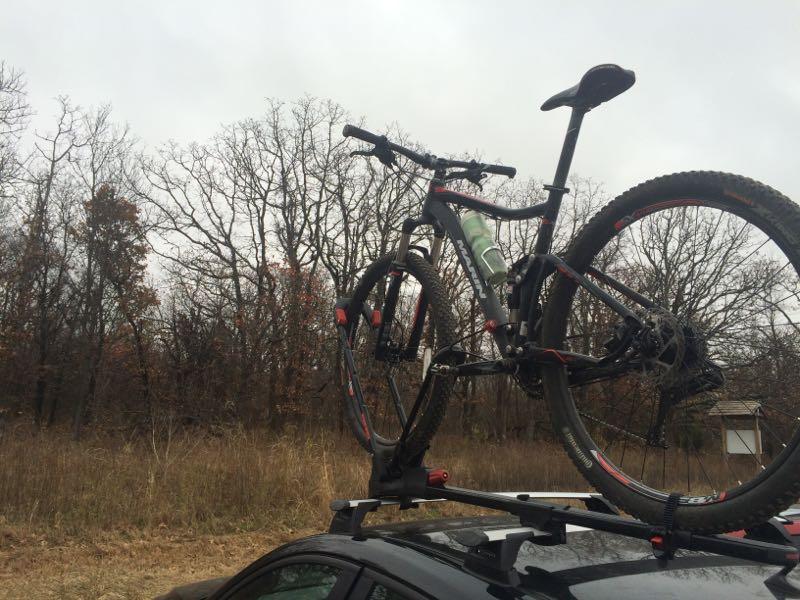 A mountain bike secured on the roof rack of a car, positioned in a natural setting with bare trees in the background under a cloudy sky. Turkey Mountain mountain bike trail.