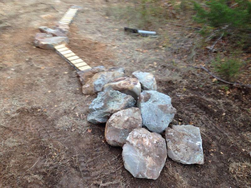 A pathway made of wooden planks is visible, winding through an outdoor area covered with dirt and grass. A cluster of large, uneven rocks is positioned alongside the pathway, adding a natural element to the landscape. In the background, sparse greenery and a faint outline of a tool can be seen. Barber Hills Trail at Pat Mayse Lake mountain bike trail.