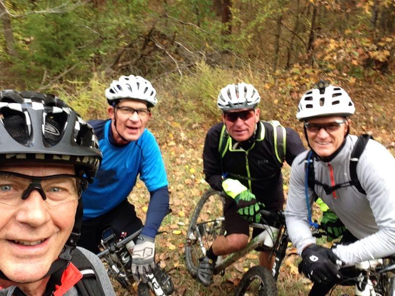 Four cyclists pose for a selfie in a forested area during autumn. Each person is wearing a helmet and cycling gear, with bicycles nearby. The background features trees and fallen leaves, indicating the season. Barber Hills Trail at Pat Mayse Lake mountain bike trail.