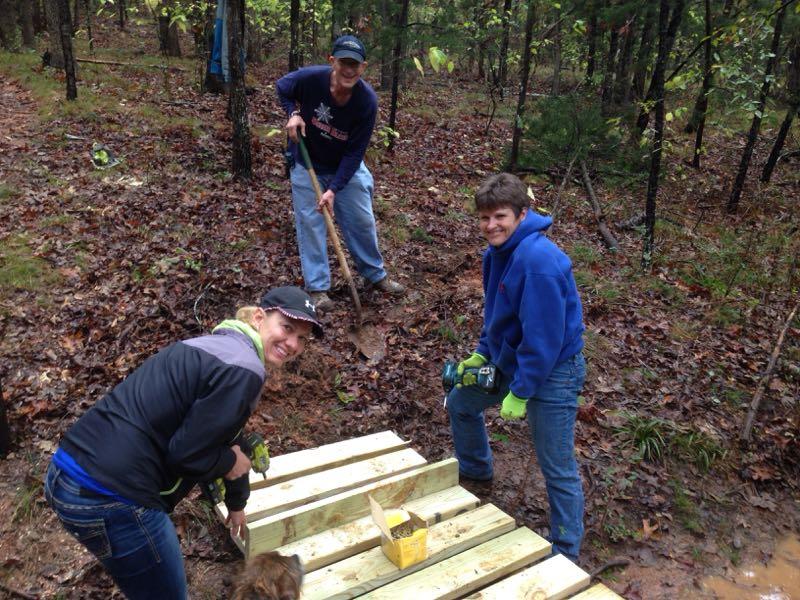 A group of three individuals is working together outdoors in a forested area. Two are smiling as they pose near some wooden planks, while one person is digging with a shovel. The ground is slightly muddy, and the scene is surrounded by trees and fallen leaves, suggesting a community project in the natural environment. Barber Hills Trail at Pat Mayse Lake mountain bike trail.
