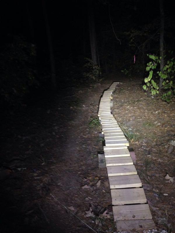 A narrow wooden path illuminated by a flashlight, leading through a dark forest with fallen leaves and sparse vegetation on either side. The path bends slightly and appears to be constructed of wooden planks resting on blocks. Surrounding trees are silhouetted against the night sky. Barber Hills Trail at Pat Mayse Lake mountain bike trail.