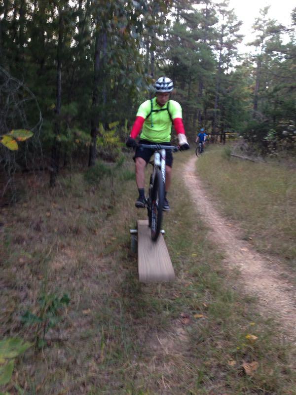 A cyclist balancing on a narrow wooden plank on a mountain bike trail, surrounded by trees and grassy terrain. Another cyclist is visible in the background, riding along the dirt path. The foreground cyclist is wearing a bright green shirt with red sleeves and a helmet. Barber Hills Trail at Pat Mayse Lake mountain bike trail.