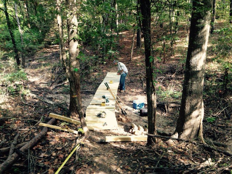 A person is working on constructing a wooden path in a forested area. Tools and materials are scattered along the trail, which is made up of wooden planks that are partially laid out. Surrounding the path are tall trees and underbrush in a natural setting. Barber Hills Trail at Pat Mayse Lake mountain bike trail.