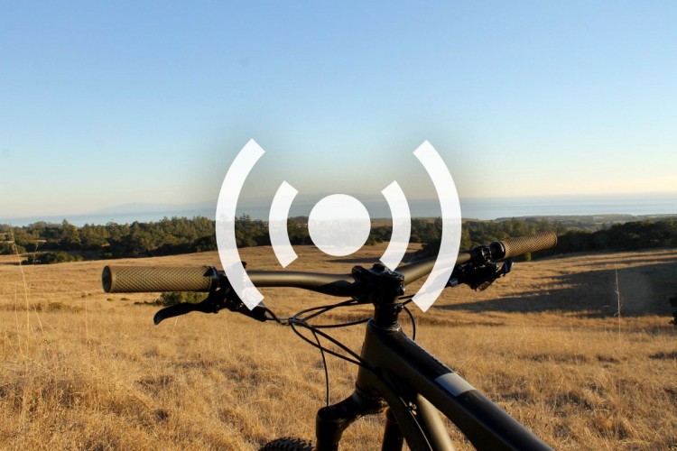 A mountain bike is positioned in the foreground with handlebars visible, overlooking a scenic landscape of rolling hills and the ocean in the background. The sky is clear with a warm, golden hue, indicating either sunrise or sunset. A graphic symbol resembling signal waves overlays the image.