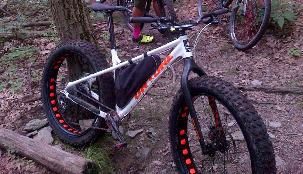 On-One Fatty: A white fat bike with bright orange accents is parked on a dirt trail surrounded by trees. The bike features wide, textured tires designed for rough terrains. In the background, other cyclists can be seen partially obscured by foliage.