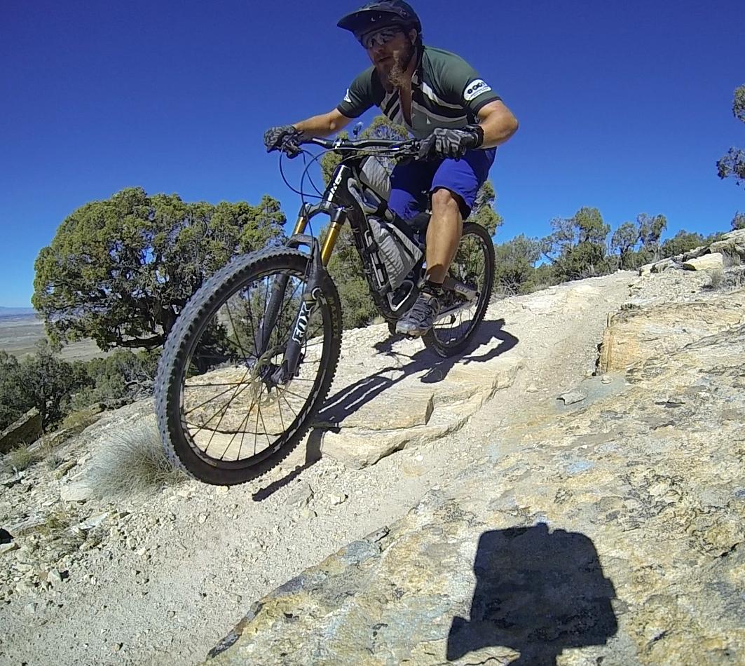 A mountain biker navigating a rocky trail under a clear blue sky. The rider is in mid-motion, demonstrating skill and balance as they ride over large stones. Green trees are visible in the background, highlighting the outdoor setting. Sidewinder mountain bike trail.