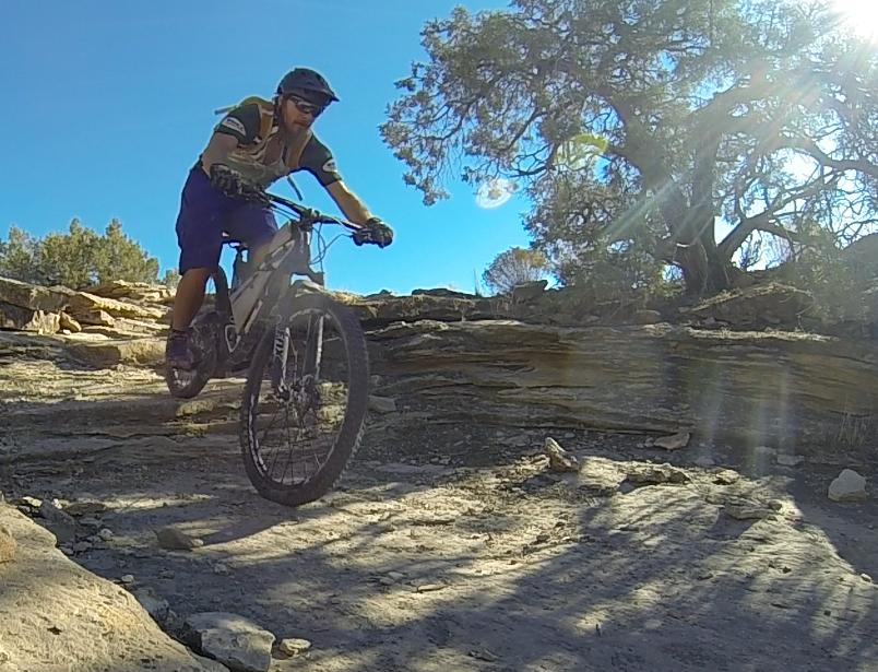 A mountain biker navigating a rocky trail under bright blue skies, with sunlight filtering through nearby trees. The rider is focused and wearing protective gear, showcasing an adventurous outdoor activity. Sidewinder mountain bike trail.