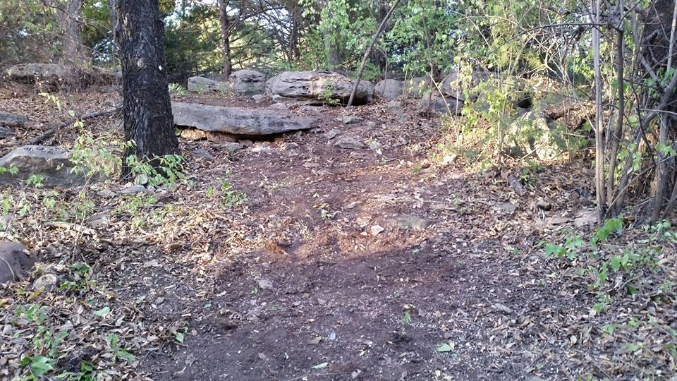 A dirt path winding through a forested area, flanked by trees and large rocks. The ground is covered in leaves and scattered stones, indicating natural terrain. Sunlight filters through the greenery, creating a serene and inviting atmosphere. Lake Ponca Trails mountain bike trail.