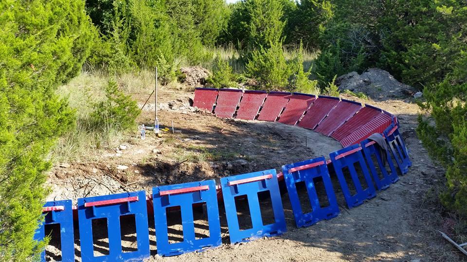 A curved arrangement of colorful barriers in a natural setting, surrounded by greenery. The barriers are blue with red tops and are placed along a dirt path that winds through the area. In the background, trees and an uneven terrain can be seen. Lake Ponca Trails mountain bike trail.