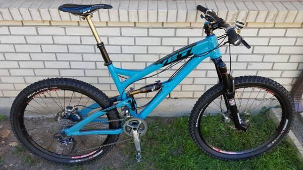 Yeti SB66: A bright blue mountain bike with a sleek design, featuring wide tires and a suspension system. The bike is positioned on grass next to a white brick wall, showcasing its detailed components including the handlebars, seat post, and gears.