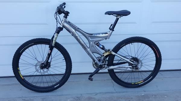 Specialized Enduro: A silver and black mountain bike leaning against a white garage door. The bike features a lightweight aluminum frame, thick knobby tires designed for off-road terrain, and front suspension forks. It has a single front chainring and adjustable seat height.